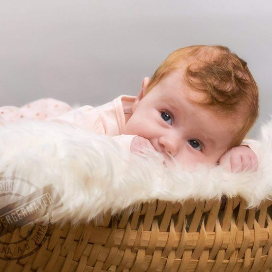 Ein Baby liegt in einem Korb auf einer weißen Pelzdecke und trägt ein rosa Outfit. Die blauen Augen des Babys blicken direkt in die Kamera, und das Baby hält seinen Kopf mit der rechten Hand.