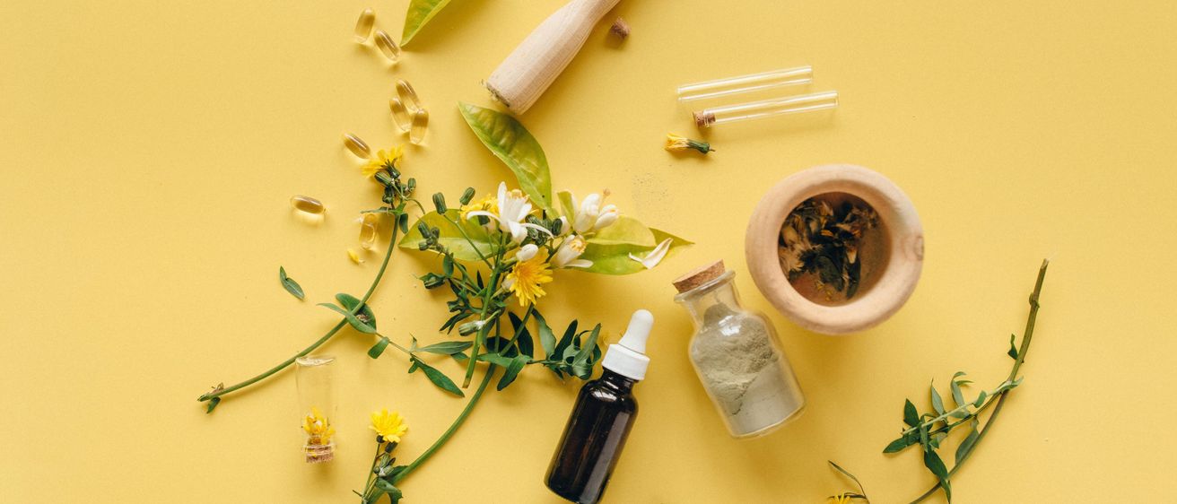 Auf einer gelben Oberfläche sind verschiedene Kräuter, ein Mörser, ein Stößel, eine Ölflasche, kleine Glasfläschchen und Blumen zu sehen.