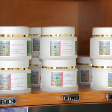 Ein Regal mit mehreren Gläsern von Hautpflegeprodukten. Die Gläser haben goldfarbene Deckel und weiße Körper. Die Gläser sind mit 'ANTIOX CREME' und 'VITAMIN-PFLEGECREME' beschriftet. Der Preis von 19,60 Euro wird auf einem digitalen Preisschild angezeigt.