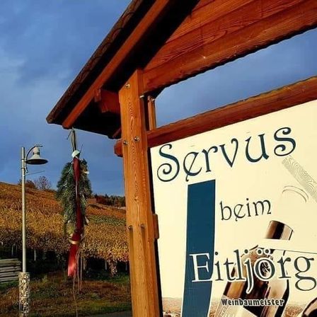 Ein Holzschild mit den Worten 'Servus beim Eitljoerg' und einem Flaschenbild ist an einem Holzrahmen befestigt. Hinter dem Schild befindet sich ein Feld mit Pflanzen und eine Straßenlampe.