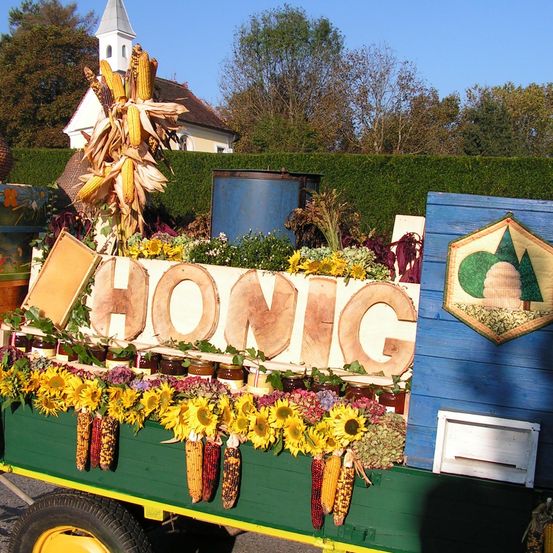 Ein grüner Anhänger gefüllt mit Sonnenblumen, Mais und einem Holzschild, auf dem HONIG steht. Dahinter befindet sich eine Kirche mit Bäumen und einem blauen Fass.