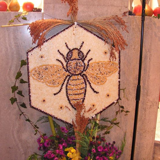 Ein hexagonales Kunstwerk, das eine Biene aus kleinen farbigen Perlen darstellt, steht prominent an einer Wand, umgeben von Blumen und Blättern.