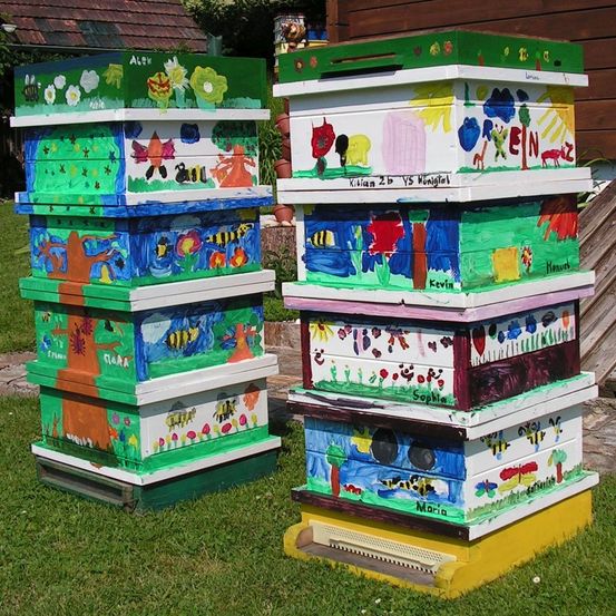 Zwei Stapel bunter Bienenstöcke sind mit verschiedenen Zeichnungen bemalt. Ein Stapel trägt Namen wie Alex, Clara und Maria. Der andere hat Namen wie Kevin, Sophia und Kilian.