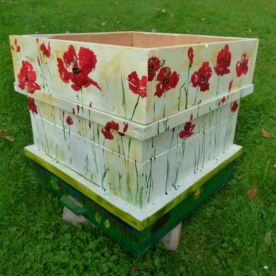 Ein hölzerner Bienenstock mit roten Blumen bemalt, auf einem Grasfeld stehend.
