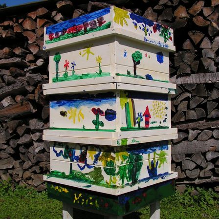 Ein Stapel farbenfroher, handbemalter Holzbeuten steht vor einem Holzhaufen. Jede Box zeigt einzigartige Kunstwerke, darunter Blumen, Bäume und ein Haus.