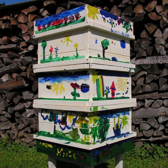 Ein Stapel farbenfroher, handbemalter Holzbeuten steht vor einem Holzhaufen. Jede Box zeigt einzigartige Kunstwerke, darunter Blumen, Bäume und ein Haus.