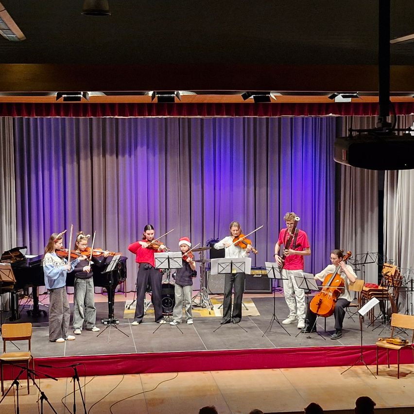 Eine Gruppe junger Musiker tritt auf der Bühne auf, beleuchtet von lila Lichtern. Geigen, ein Cello und ein Klavier sind sichtbar. Sie sind in Freizeitkleidung gekleidet, einige tragen Weihnachtsmützen.