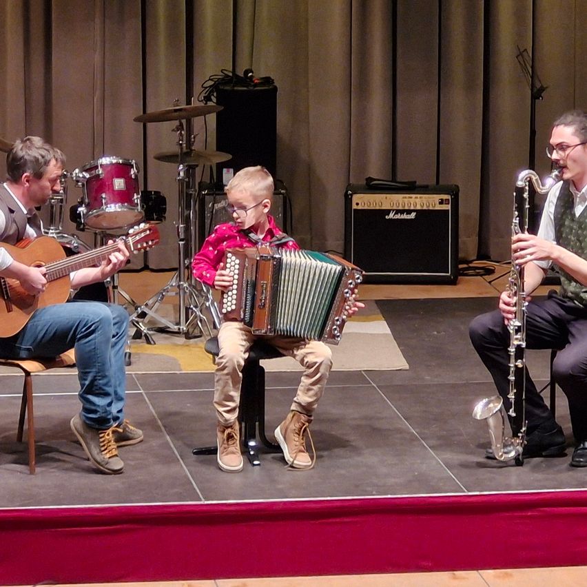 Drei Musiker spielen auf einer Bühne. Ein Mann spielt Gitarre, ein junger Junge spielt Akkordeon und ein weiterer Mann spielt Saxophon. Hinter ihnen stehen ein Schlagzeug und ein Lautsprecher.
