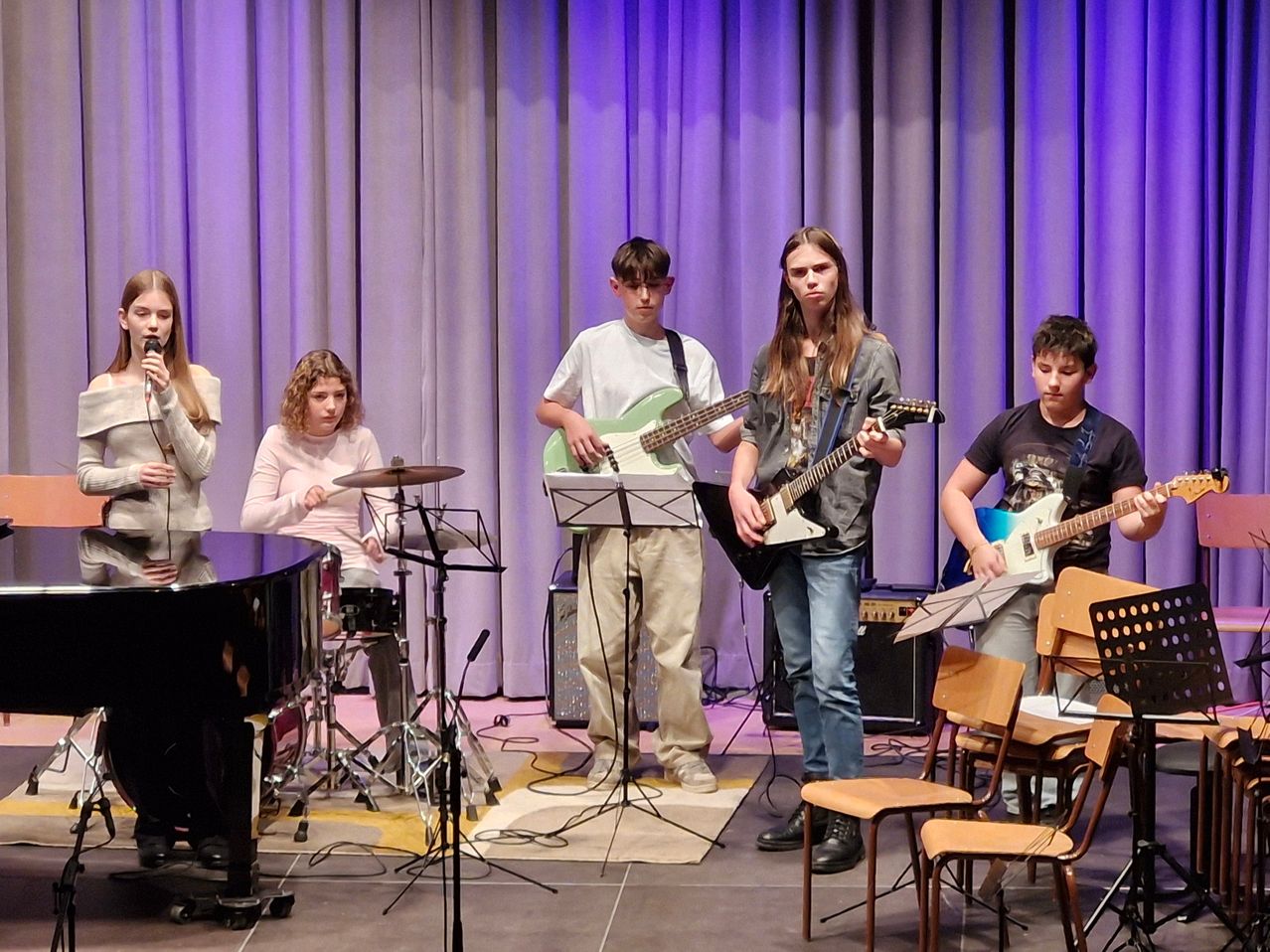 Eine Gruppe von Kindern tritt auf der Bühne auf. Einer spielt Klavier und die anderen Gitarren und Schlagzeug. Sie stehen auf einem Teppich, mit einem lila Vorhang im Hintergrund.