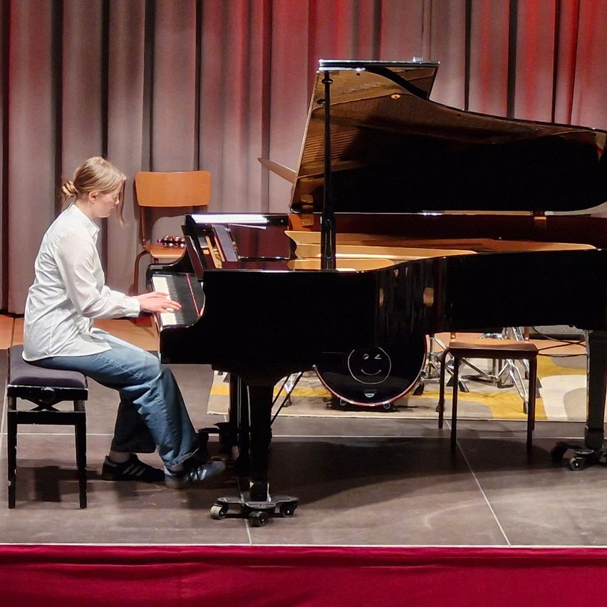 Eine junge Frau spielt Klavier auf einer Bühne mit roten Vorhängen im Hintergrund. Sie trägt ein weißes Hemd und blaue Jeans.