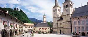Ein Kirchenplatz mit spazieren gehenden Menschen, umgeben von Gebäuden und einem Brunnen.