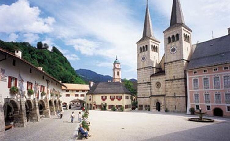 Ein Kirchenplatz mit spazieren gehenden Menschen, umgeben von Gebäuden und einem Brunnen.