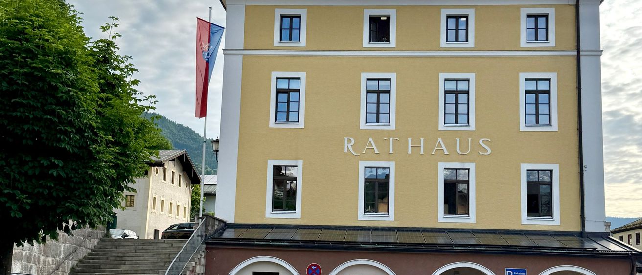 Ein gelbes Gebäude mit der Aufschrift Rathaus steht vor einem Berg. Es hat viele Fenster und eine Fahne. Ein schwarzes Auto ist auf der linken Seite geparkt.