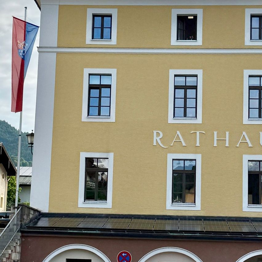 Ein gelbes Gebäude mit der Aufschrift Rathaus steht vor einem Berg. Es hat viele Fenster und eine Fahne. Ein schwarzes Auto ist auf der linken Seite geparkt.