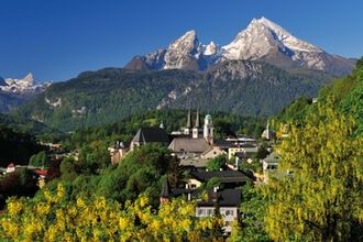Ein malerisches Dorf am Fuße eines Gebirgszugs mit schneebedeckten Gipfeln, umgeben von üppigen grünen Bäumen und gelben Blumen unter einem klaren blauen Himmel.