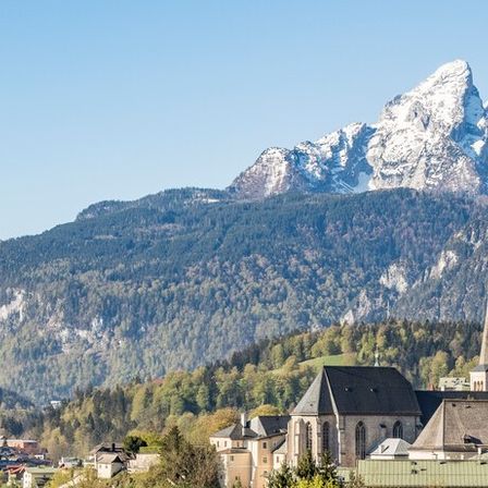 Ein kleines Städtchen mit Gebäuden, Bäumen und einer Kirche liegt in den Alpen, mit schneebedeckten Bergen im Hintergrund.