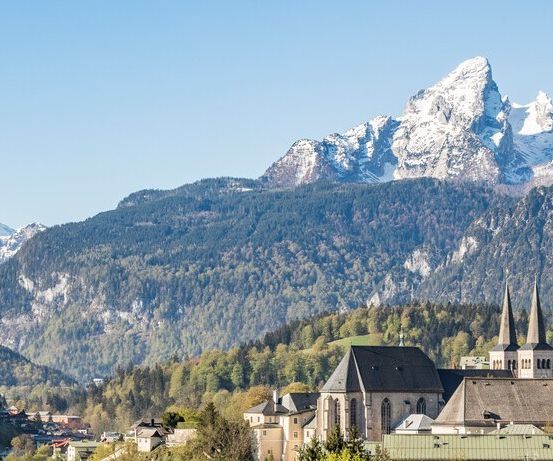 Ein kleines Städtchen mit Gebäuden, Bäumen und einer Kirche liegt in den Alpen, mit schneebedeckten Bergen im Hintergrund.