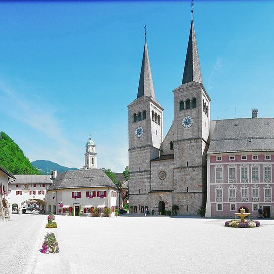 Eine Kirche mit zwei Türmen und Uhren steht vor einem Stadtplatz mit einem Brunnen. Gebäude und Grünflächen umgeben die Kirche. Im Hintergrund sind Berge sichtbar.