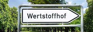 Ein Metallschild mit einem weißen Pfeil nach rechts, der das Wort 'Wertstoffhof' in Schwarz anzeigt. Das Schild ist auf einem Pfosten in einem grünen Bereich montiert, mit mehreren Bäumen im Hintergrund.