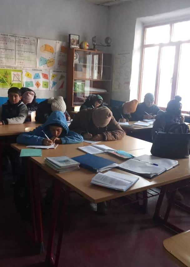 Ein Klassenzimmer mit mehreren Schülern, die Mützen und Schals tragen. Sie sitzen an Holztischen und schreiben mit Stiften auf Papier. Auf den Tischen liegen Bücher, Notizbücher und eine Handtasche. Im Hintergrund steht ein Regal mit verschiedenen Gegenständen.