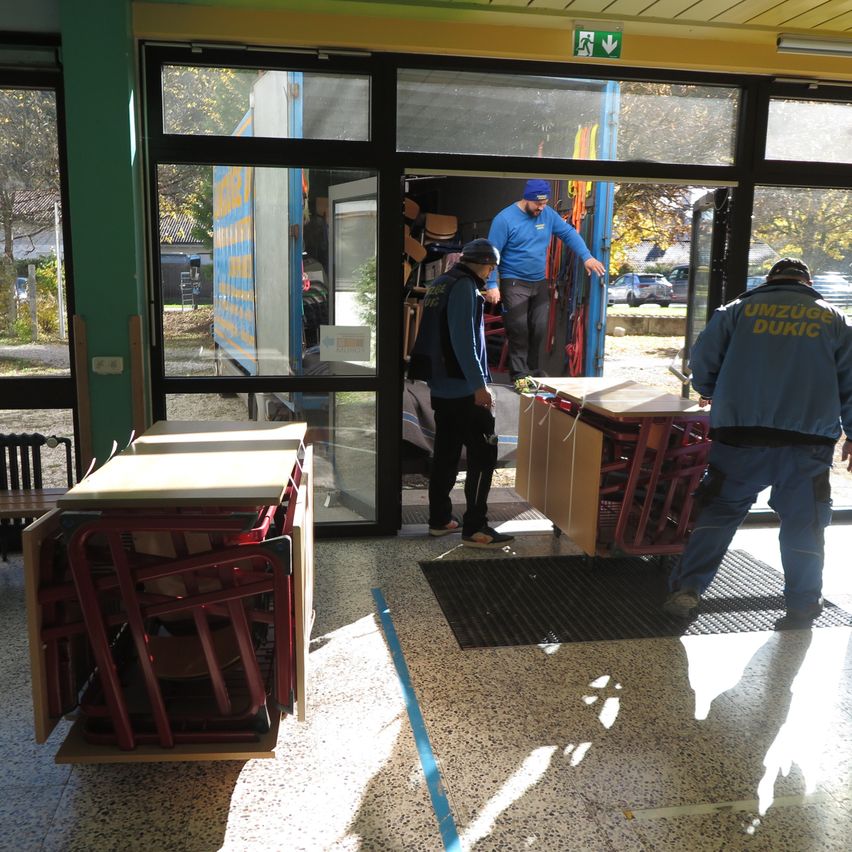 Zwei Arbeiter entladen einen Lastwagen mit Stühlen und Tischen in einem hell erleuchteten Raum. Eine Person an der Tür hilft, während andere zusehen.