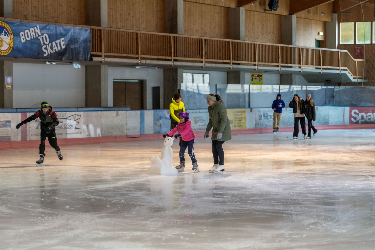 Eine Eisbahn, auf der eine Frau und ein Kind mit einem Schneemann Schlittschuh laufen, während andere in der Hintergrund laufen.