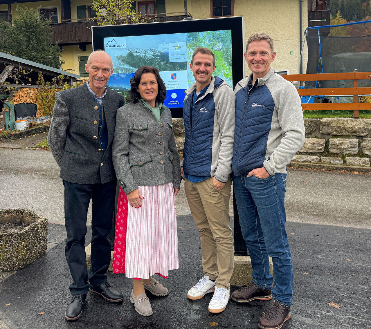 Vier Personen stehen zusammen vor einem großen Informationsschild. Sie lächeln und scheinen für ein Foto zu posieren. Der Mann links trägt ein traditionelles Outfit, während die anderen lässig gekleidet sind. Im Hintergrund befindet sich ein Gebäude mit Balkonen und einem Trampolin.