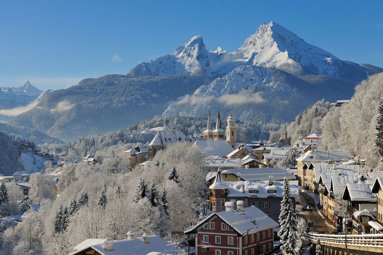 Schneebedeckte Stadt in den Alpen, mit Türmen und Giebeln, die durch den Schnee ragen, unter einem blauen Himmel.