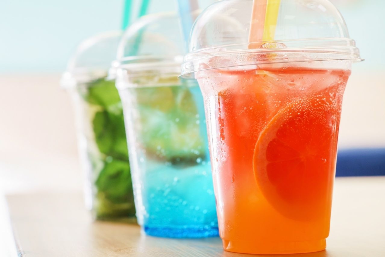 Drei Plastikbecher mit Deckeln und Strohhalmen, die orange, grüne und blaue Getränke auf einem Holztisch enthalten.