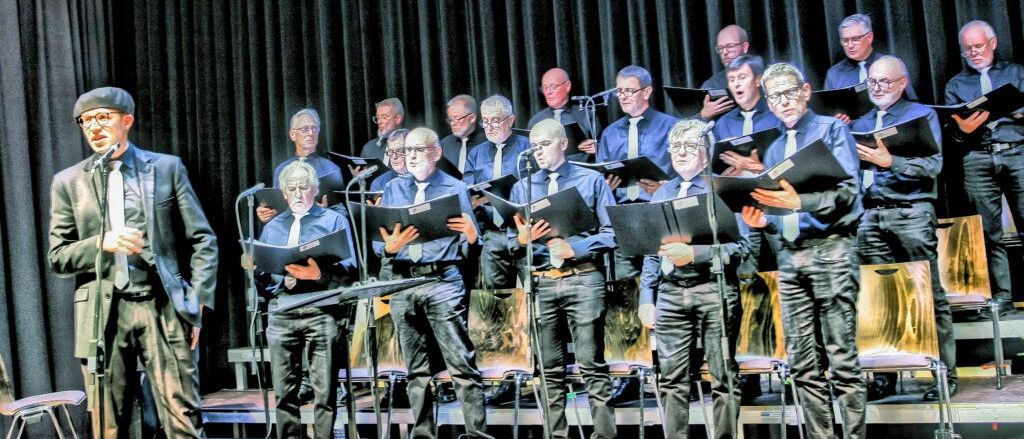 Ein Chor älterer Männer in formeller Kleidung tritt auf der Bühne auf, mit Mikrofonen und Notenständern vor ihnen. Dahinter dient ein schwarzer Vorhang als Hintergrund.