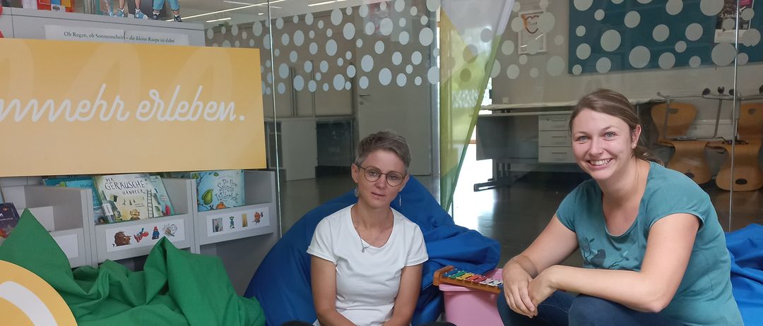 Zwei Frauen sitzen auf einem gelben Sitzsack mit Musikinstrumenten, ein Banner mit der Aufschrift 'mehr erleben' im Hintergrund.