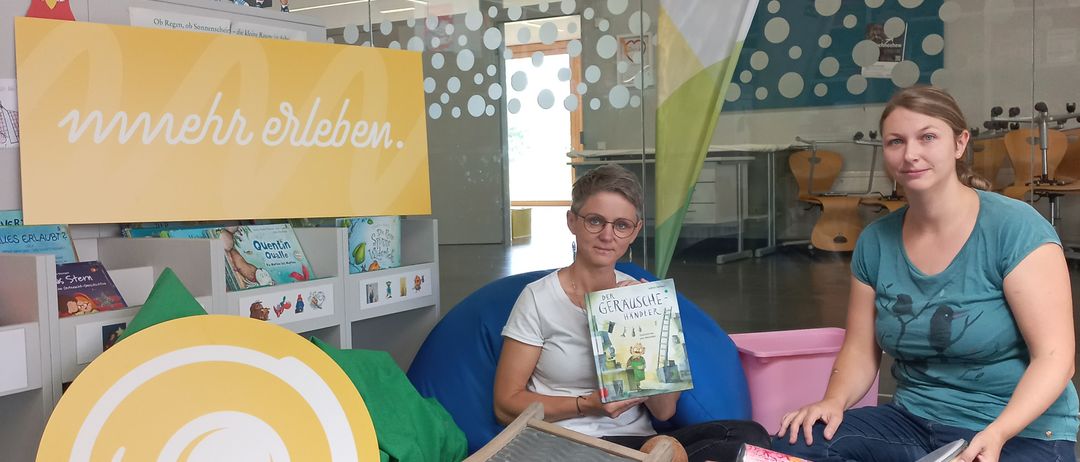 Zwei Frauen sitzen in einer Bibliothek, umgeben von Kinderbüchern und Spielzeug. Eine hält ein Buch, und beide lächeln für die Kamera. Ein buntes Schild steht 'mehr erleben'.