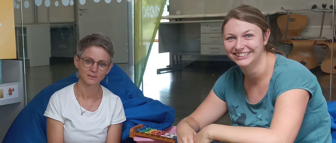 Zwei Frauen sitzen auf einem gelben Beanbag-Sessel mit Musikinstrumenten vor ihnen. Sie lächeln und posieren für ein Foto.