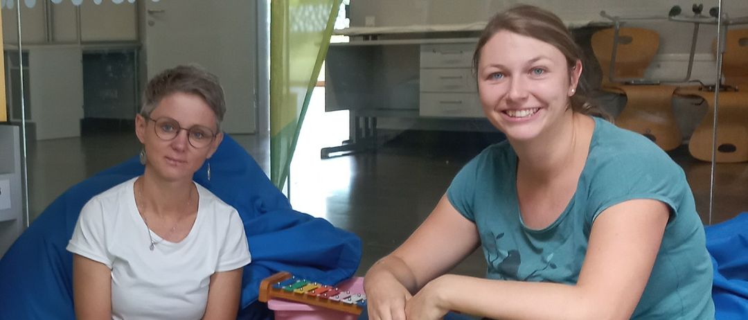 Zwei Frauen sitzen auf Bohnensäcken in einem Raum mit Tupfenwänden und einem weißen Banner mit der Aufschrift BibliooTherapy. Sie lächeln für ein Foto.