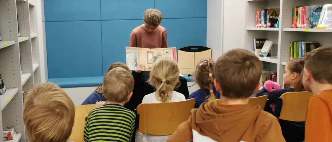 Eine Gruppe von Kindern sitzt in Stühlen in einer Bibliothek und hört einer Frau zu, die ein Buch liest.