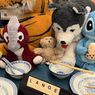 Ein Tiger, ein Panda und verschiedene andere Kuscheltiere stehen auf einem Tisch mit blauen Tellern, Schüsseln, Chopsticks und einem Schild, das 'Lange Nacht' besagt. Dahinter sind Bücher in Regalen ausgestellt.