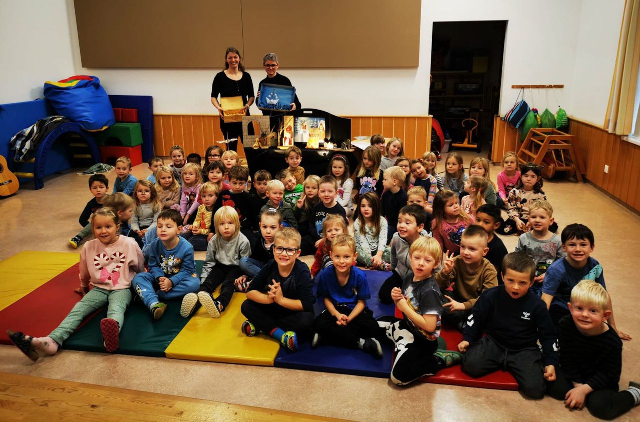 Eine Gruppe von Kindern sitzt auf farbenfrohen Matten auf dem Boden. Ein Mann und eine Frau stehen hinter einem Tisch mit einer Krippe.
