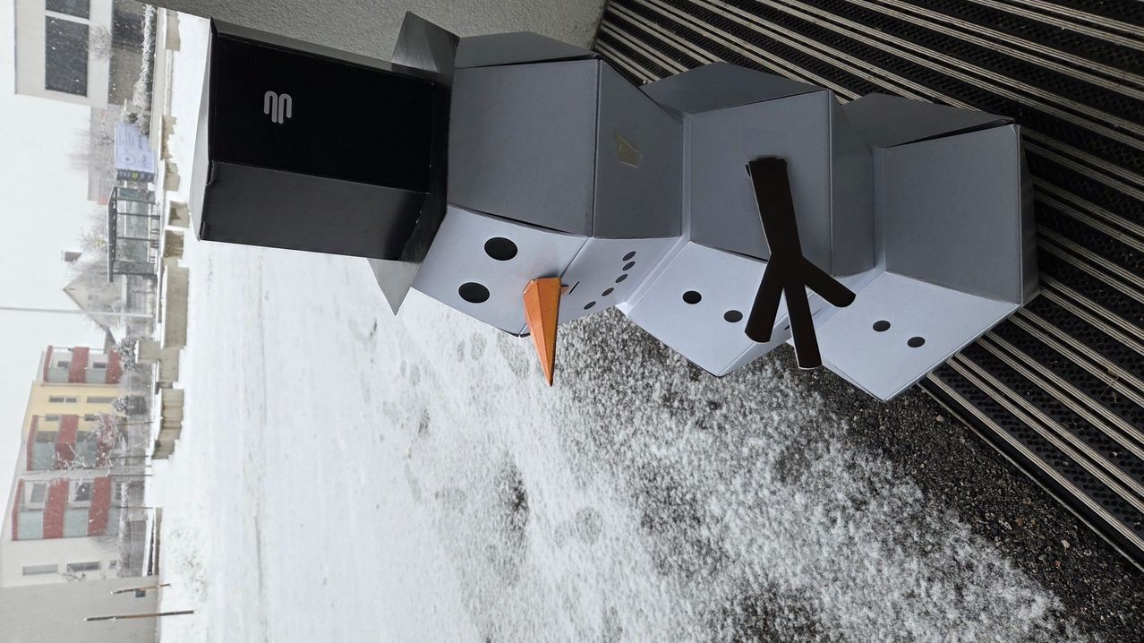 Ein Schneemann aus Kartons, mit einer Karottennase und Augen, sitzt auf einer verschneiten Oberfläche. Ein schwarzer Kasten mit einem weißen 'N' befindet sich dahinter.
