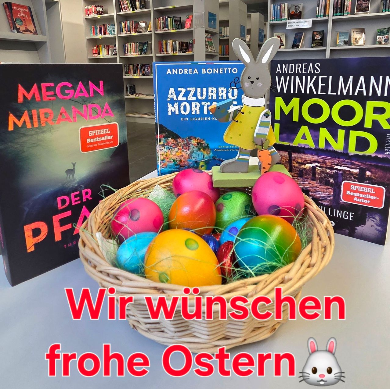 Ein Korb voller bunter Ostereier steht auf einem Tisch vor dem Hintergrund von Büchern. Der deutsche Text lautet 'Wir wuenschen frohe Ostern' und ein Häschen ist unten rechts.