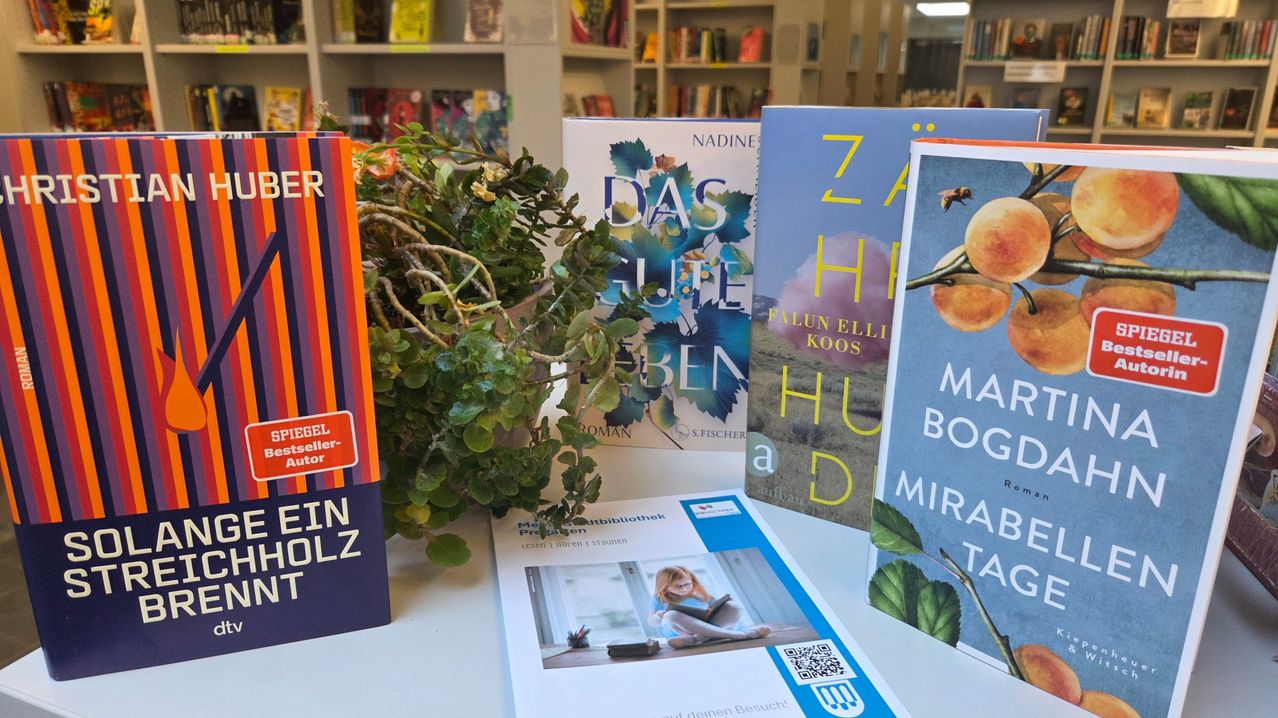 Bücher und eine Pflanze auf einem weißen Tisch, mit einer Bibliothek im Hintergrund. Die Titel sind "Das Gute Leben" und "Zahira." Eine Frau liest im Bild.