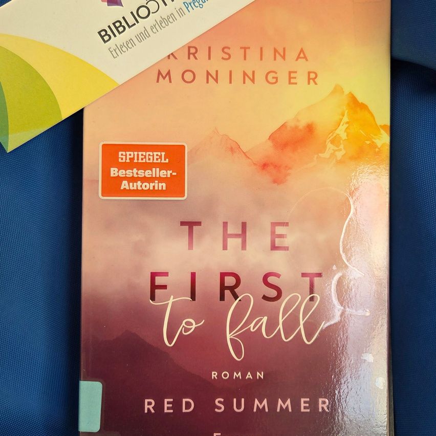 A book titled 'The First to Fall' by Kristina Moninger, a bestseller author, is placed on a blue surface. The cover features a watercolor mountain landscape.