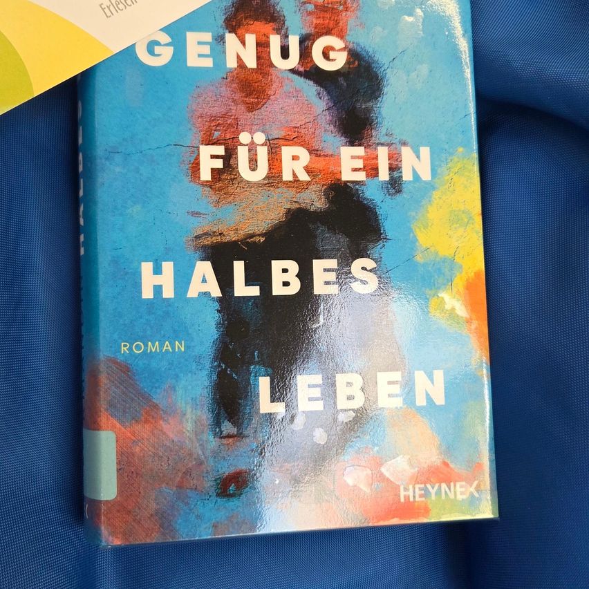 A book titled 'Genug für ein halbes Leben' by Heynex, featuring a colorful abstract painting on the cover, is placed on a blue surface.