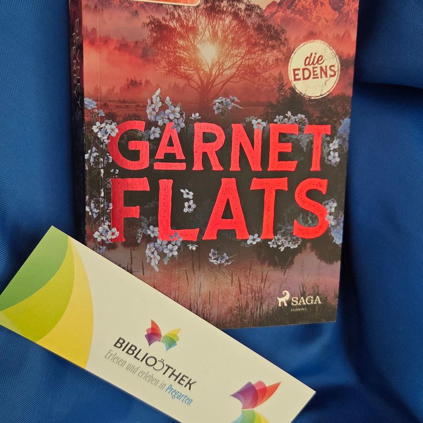 A book titled 'Garnet Flats' by Die Edens is placed on a blue surface. The cover features a sunset with a silhouette of a tree and flowers.