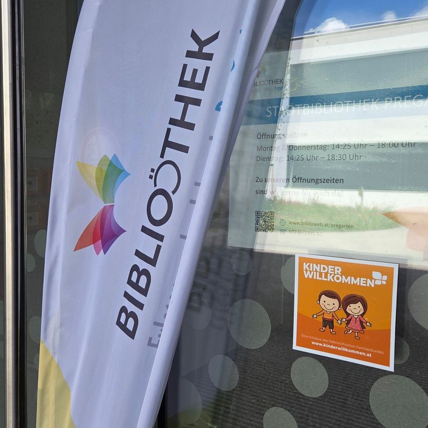 Ein weißer Banner mit dem Text 'BIBLIOTHEK' wird auf einem Glasfenster angezeigt. Das Fenster hat auch einen bunten Aufkleber mit dem Text 'KINDER WILLKOMMEN' und einem Bild von zwei Kindern. Der Aufkleber ist auf dem Glasfenster angebracht. Das Glasfenster spiegelt den Himmel und die Umgebung wider.