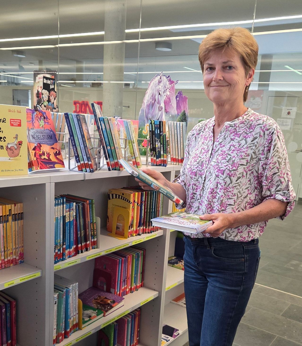 Eine Frau mit kurzen Haaren steht vor einem Bücherregal und hält zwei Bücher in den Händen. Hinter ihr spiegelt eine Glaswand einen Raum mit Bücherregalen, einem Pferdebild und einem Banner wider.