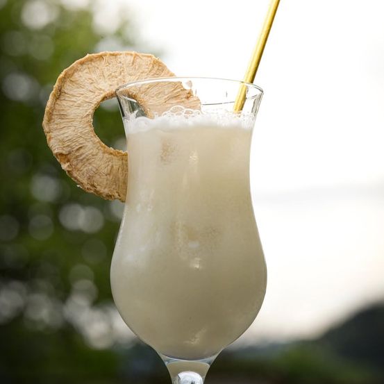 Ein Nahaufnahme eines cremigen Cocktails in einem hohen Glas mit einem goldenen Strohhalm und einer Ananasscheibe obenauf.