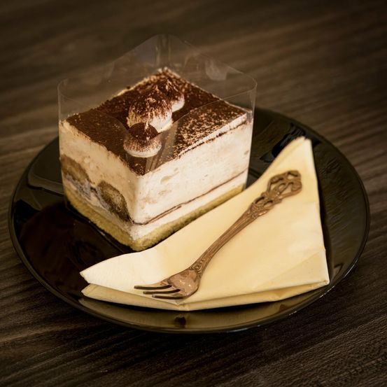 Ein Stück Tiramisu-Dessert wird auf einem schwarzen Teller präsentiert, bedeckt mit einer durchsichtigen Plastikfolie und begleitet von einer silbernen Gabel auf einem gefalteten weißen Serviettentuch.