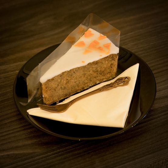 Ein Stück Karottenkuchen mit weißer Glasur und Orangenschale auf einem schwarzen Teller, bedeckt mit durchsichtigem Plastik und begleitet von einer silbernen Gabel auf einem Serviettentuch.