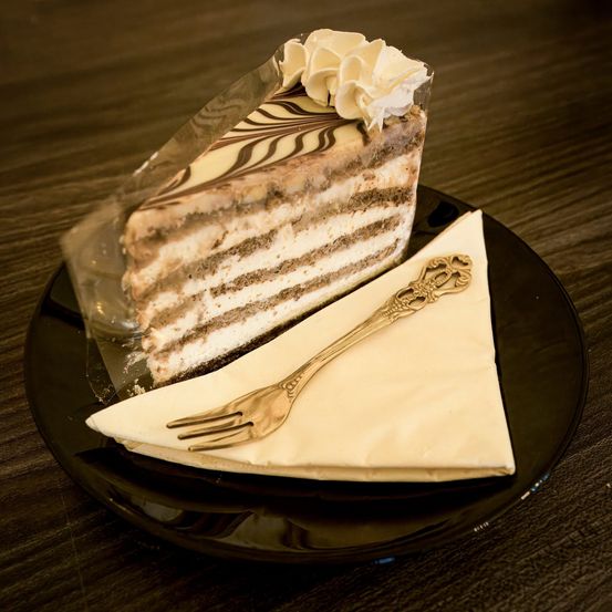 Ein Stück Schichtkuchen mit Schokolade und Sahne auf einem schwarzen Teller, begleitet von einer goldenen Gabel auf einem weißen Serviettentuch.