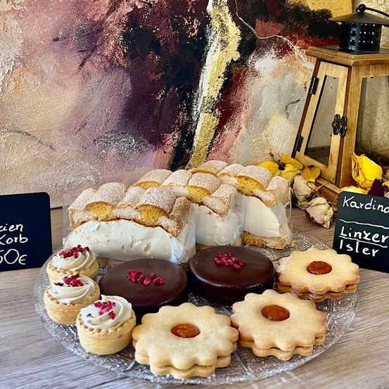 Eine Glasplatte mit verschiedenen Desserts wie Schokoladenkuchen, Törtchen und Keksen auf einem Holztisch vor dem Hintergrund eines abstrakten Gemäldes und einer Laterne.